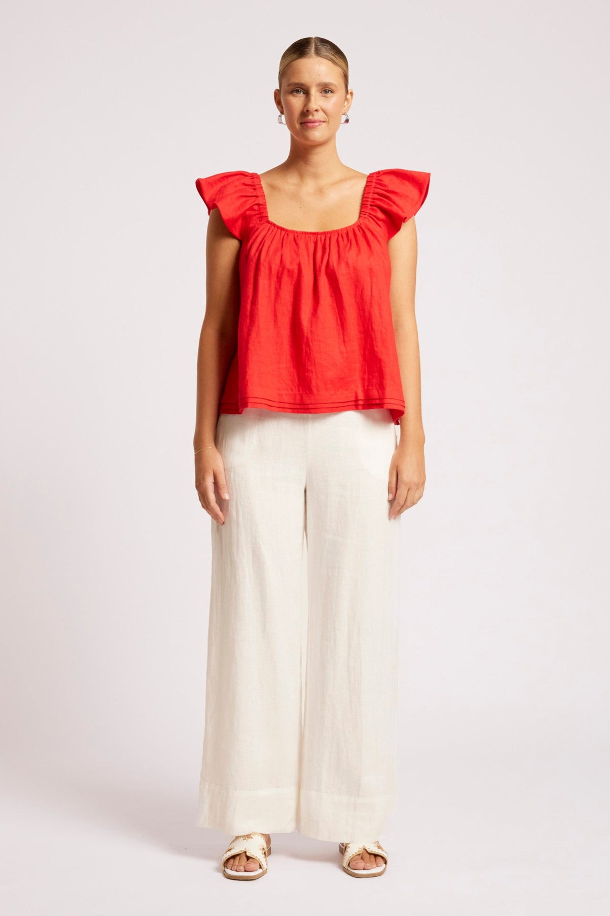 A woman modeling a bright red ruffled-sleeve crop top with a square neckline, paired with off-white wide-leg trousers and beige sandals, against a plain light gray studio background.