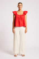 A woman modeling a bright red ruffled-sleeve crop top with a square neckline, paired with off-white wide-leg trousers and beige sandals, against a plain light gray studio background.