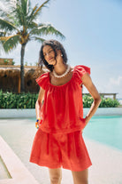 A woman wearing a bright red two-piece lounge set with ruffled sleeves and matching shorts, standing by a swimming pool with a palm tree and resort background.