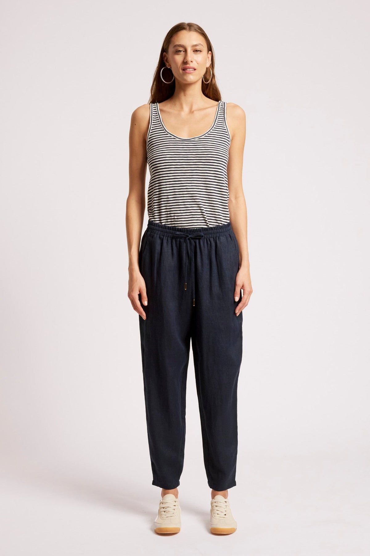 A woman modeling a sleeveless black and white striped tank top paired with dark navy drawstring pants and beige sneakers, standing against a light gray studio background.