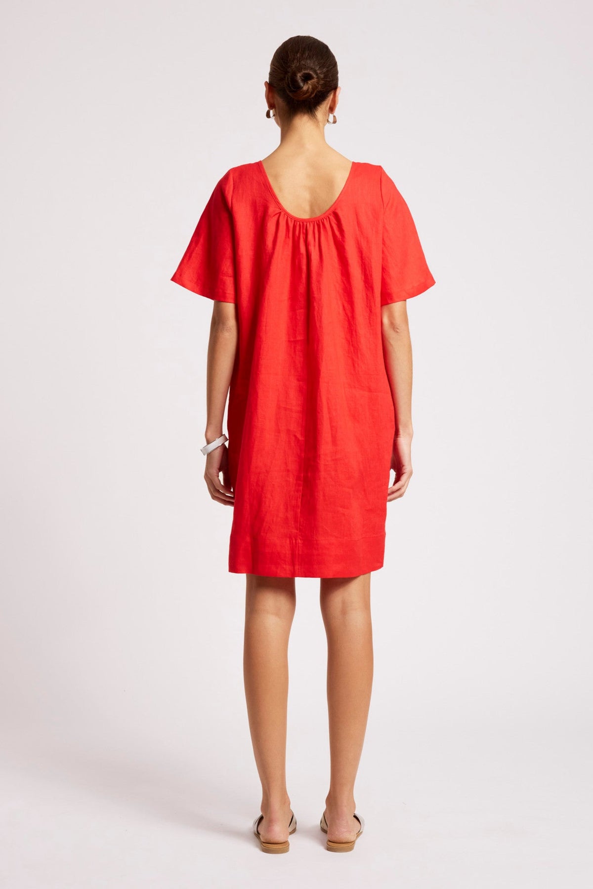 A woman viewed from behind wearing a bright red short-sleeve dress with a gathered neckline, standing against a plain light gray studio background.