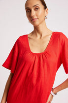 A woman wearing a bright red short-sleeve top with a wide scoop neckline and gathered detailing at the chest, photographed against a plain light background.