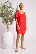 A woman modeling a bright red short-sleeve linen dress with a gathered neckline, standing next to a potted olive tree against a light gray studio background.
