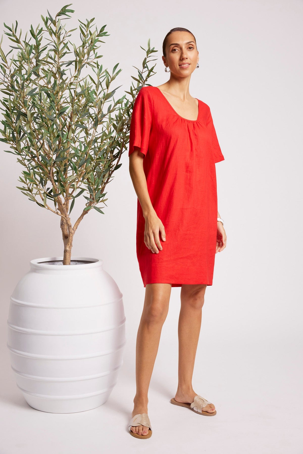A woman modeling a bright red short-sleeve linen dress with a gathered neckline, standing next to a potted olive tree against a light gray studio background.