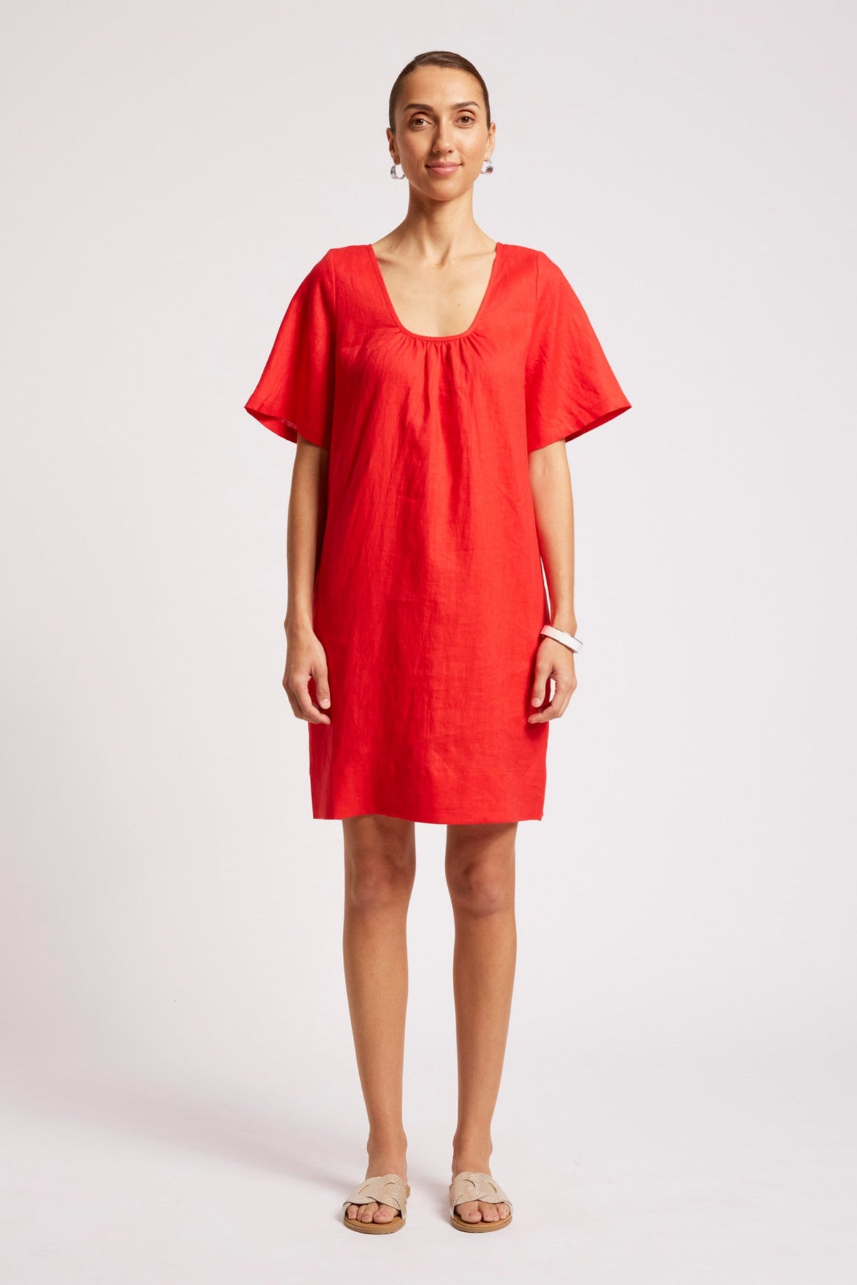 A woman modeling a bright red linen dress with short flutter sleeves, a square neckline with gathered detailing, and a relaxed fit, standing against a light gray studio background.