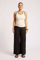 A woman modeling a cream ribbed tank top with black trim and black wide-leg pants against a light gray studio background.