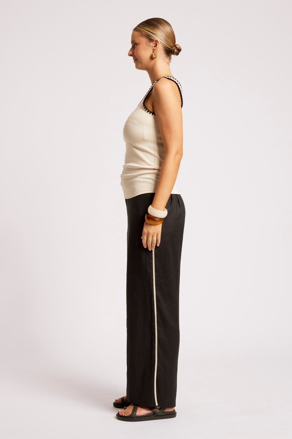 A woman shown in profile wearing a cream ribbed knit tank top with black beaded trim and black wide-leg pants with a white side stripe, standing against a light gray studio background.