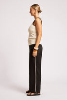 A woman shown in profile wearing a cream ribbed knit tank top with black beaded trim and black wide-leg pants with a white side stripe, standing against a light gray studio background.