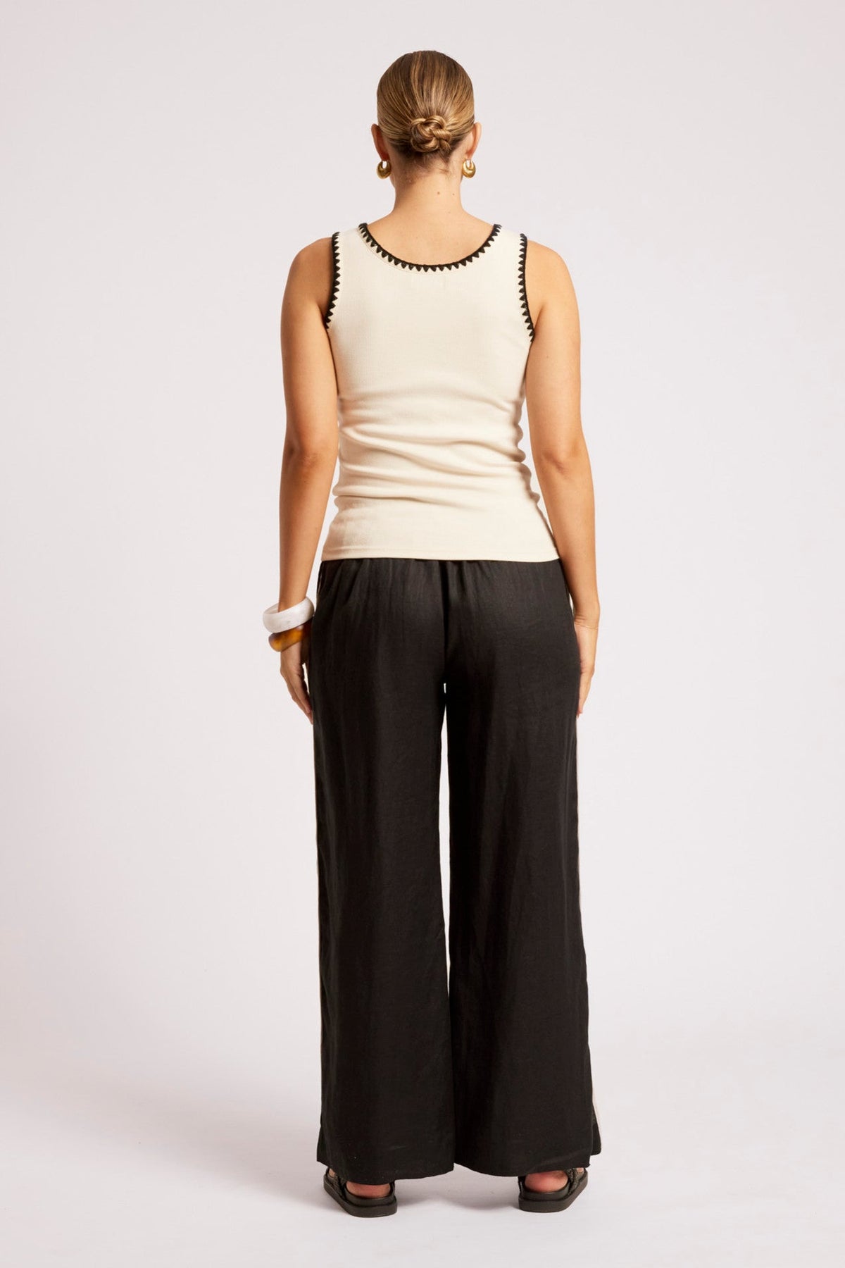 A woman shown from the back wearing a cream ribbed tank top with black trim and black wide-leg pants, standing against a light gray studio background.