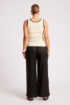 A woman shown from the back wearing a cream ribbed tank top with black trim and black wide-leg pants, standing against a light gray studio background.
