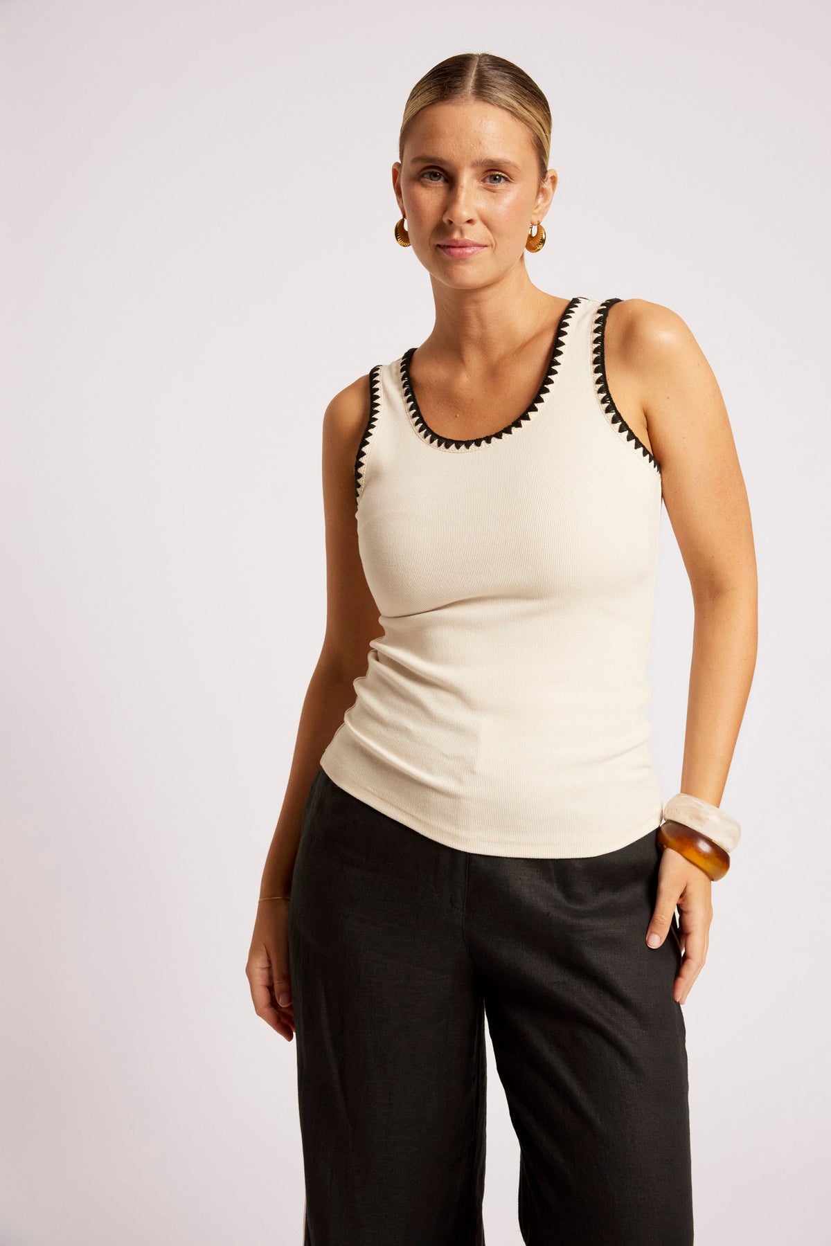 A woman modeling a cream ribbed knit tank top with black crochet trim along the scoop neckline, paired with black pants, against a light gray studio background.