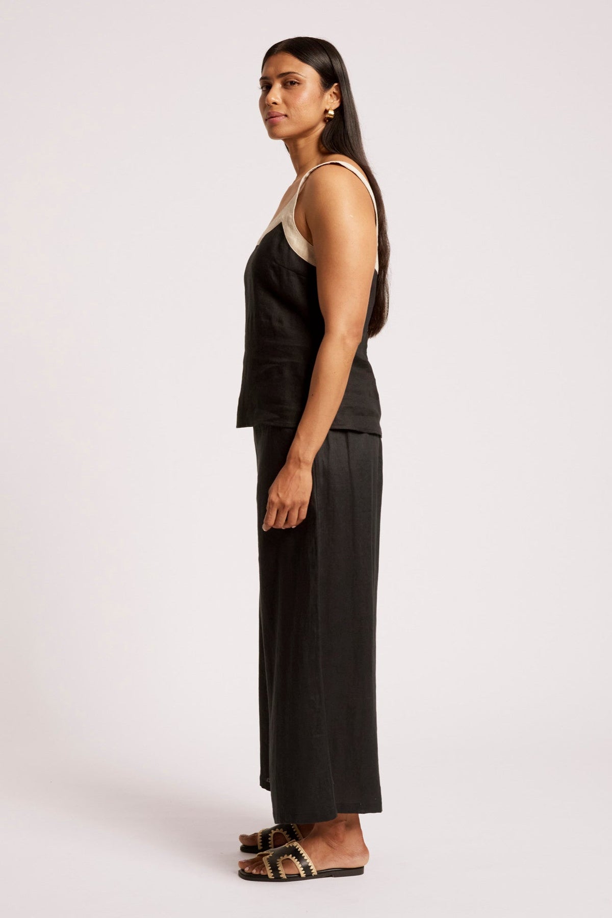 A woman stands in profile wearing a black sleeveless top and matching wide-leg pants, paired with brown strappy flat sandals, against a plain light gray studio background.