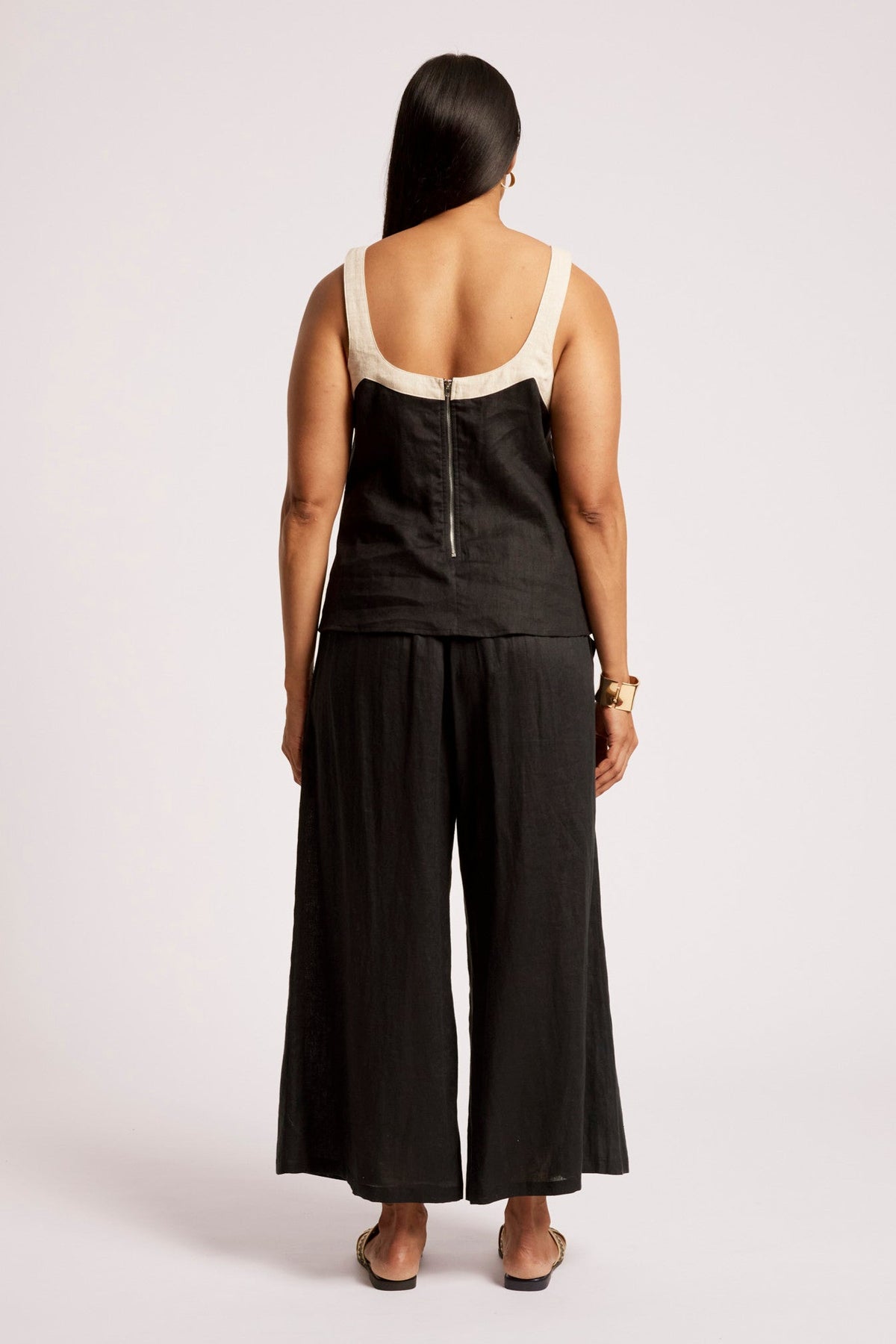 A woman viewed from the back wearing a black sleeveless jumpsuit with cream-colored shoulder straps and a matching black wide-leg pant leg, standing against a light gray studio background.
