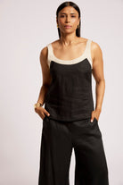 A woman modeling a black linen tank top with contrasting beige trim and black wide-leg pants against a light gray studio background.