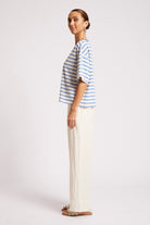 A woman modeling a short-sleeve striped top and wide-leg trousers in a studio setting. The top features thin blue and white horizontal stripes, paired with off-white wide-leg pants and brown sandals.