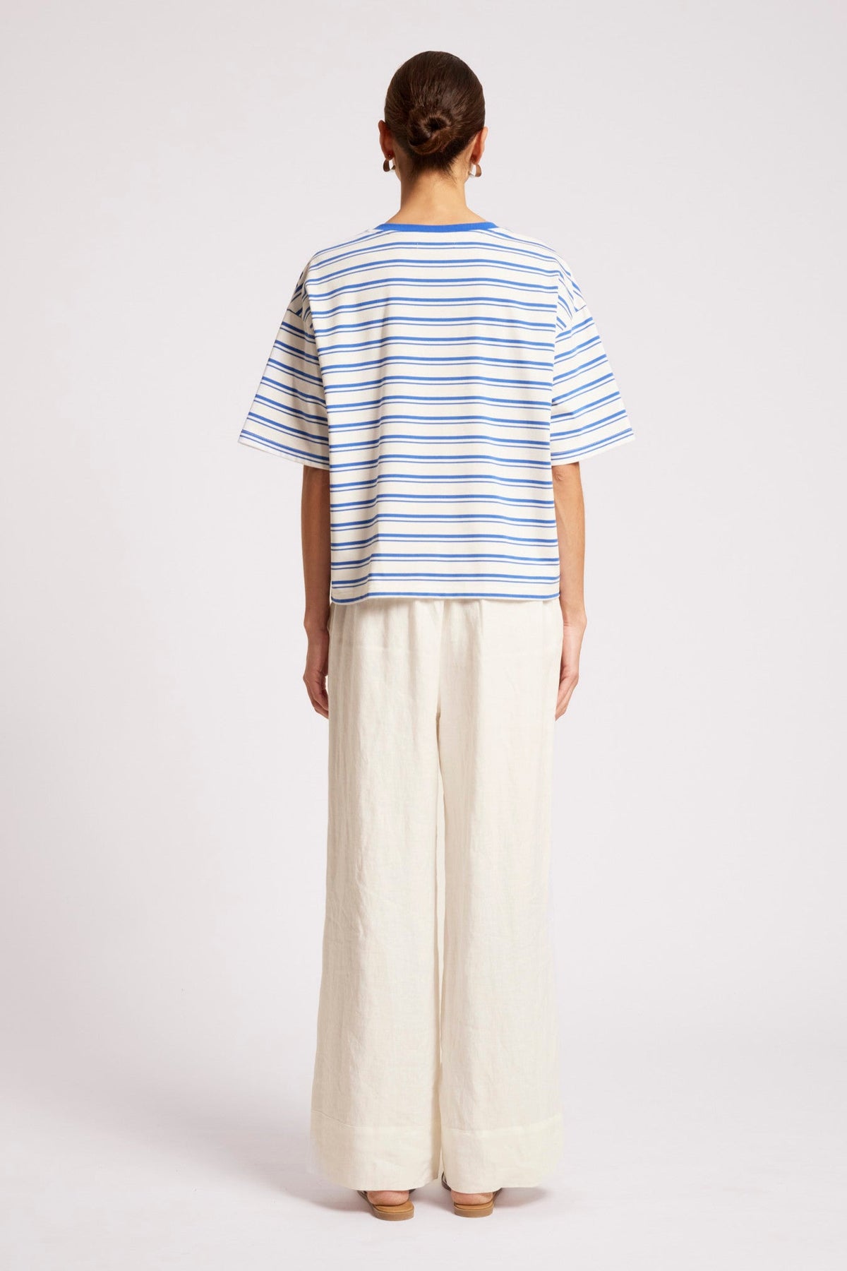 A model shown from the back wearing a white short-sleeve t-shirt with thin blue horizontal stripes and off-white wide-leg trousers, set against a plain light gray studio background.