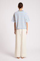 A model shown from the back wearing a white short-sleeve t-shirt with thin blue horizontal stripes and off-white wide-leg trousers, set against a plain light gray studio background.