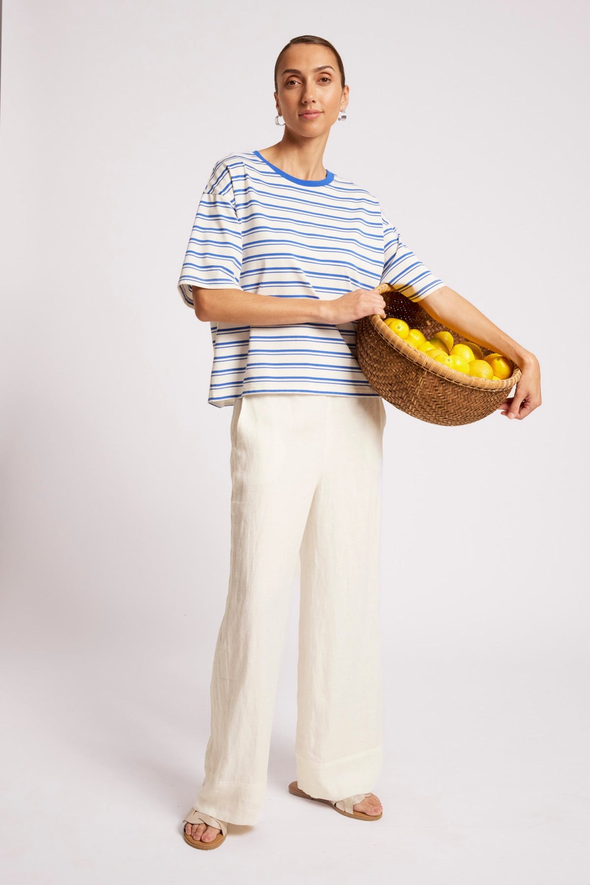 A woman modeling a blue and white striped short-sleeve top with cream wide-leg trousers, holding a woven basket of lemons against a light gray studio background.