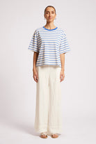 A woman modeling a short-sleeve striped t-shirt with blue and white horizontal stripes and a blue crew neck, paired with wide-leg cream trousers, against a light gray studio background.