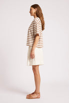A woman in side profile wearing a short-sleeve striped top with brown and beige horizontal stripes, a cream-colored mini skirt, and tan flat sandals, set against a plain light gray studio background.