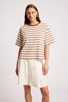 A woman modeling a short-sleeve crewneck top with thin brown and beige horizontal stripes, paired with off-white shorts, against a light gray studio background.