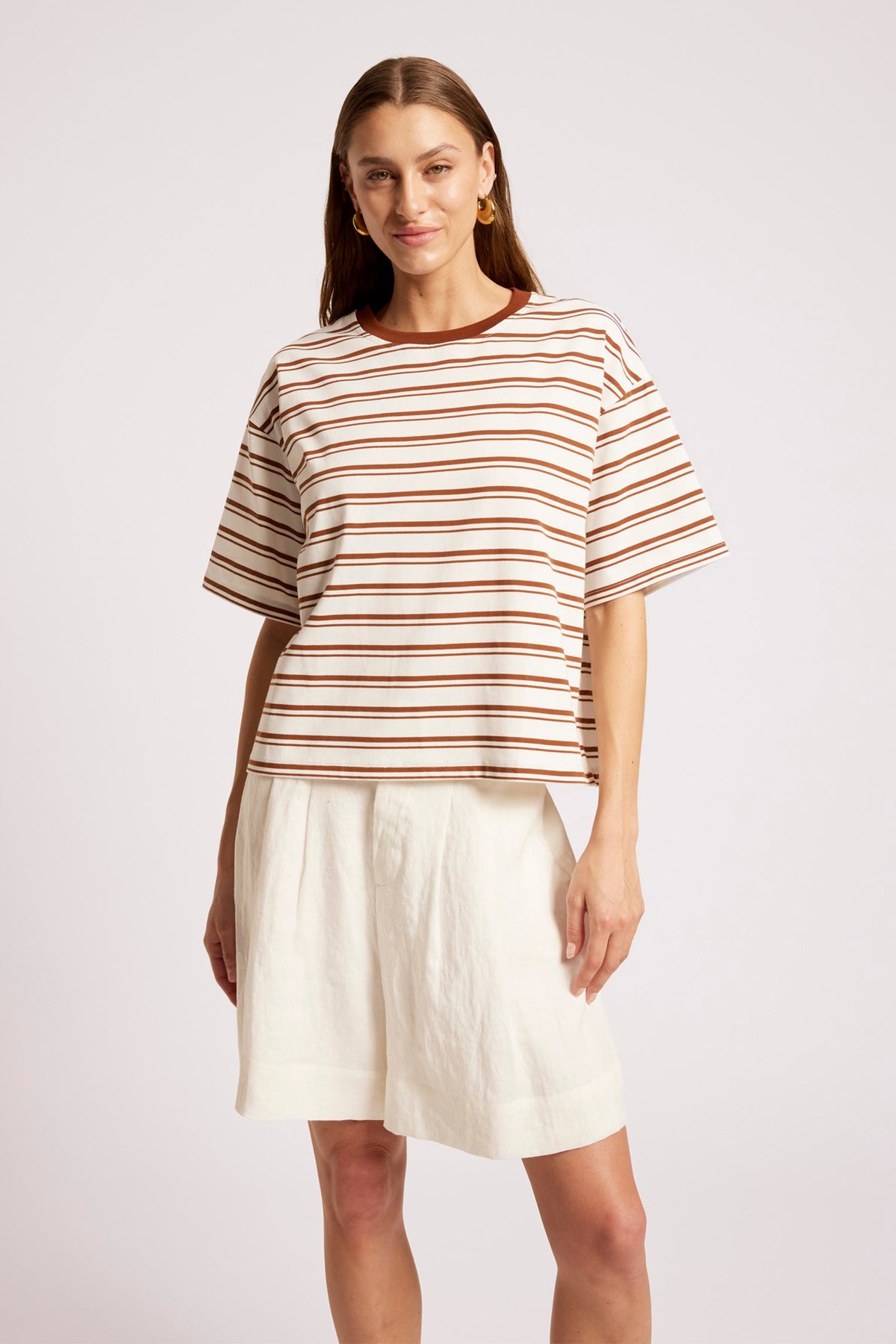 A woman modeling a short-sleeve crewneck top with thin brown and beige horizontal stripes, paired with off-white shorts, against a light gray studio background.