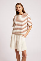 A woman modeling a short-sleeve crewneck top with thin brown and beige horizontal stripes, paired with an off-white linen skirt, against a light gray studio background.