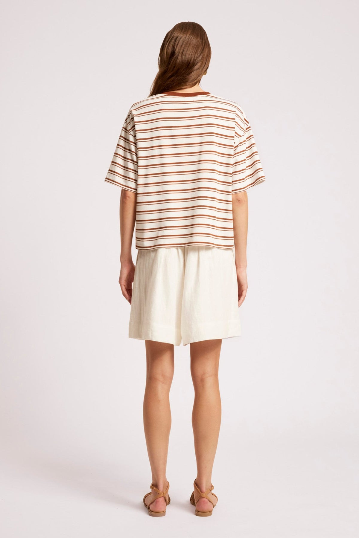 A woman shown from the back wearing a short-sleeve striped t-shirt in beige and brown with cream shorts, standing against a light gray studio background.