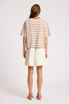 A woman shown from the back wearing a short-sleeve striped t-shirt in beige and brown with cream shorts, standing against a light gray studio background.