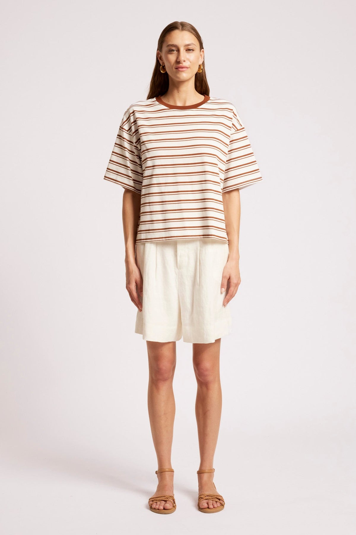 A woman modeling a short-sleeve crewneck top with thin brown and beige horizontal stripes, paired with off-white shorts and tan sandals, against a light gray studio background.