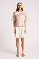 A woman modeling a short-sleeve crewneck top with thin brown and beige horizontal stripes, paired with off-white shorts and tan sandals, against a light gray studio background.