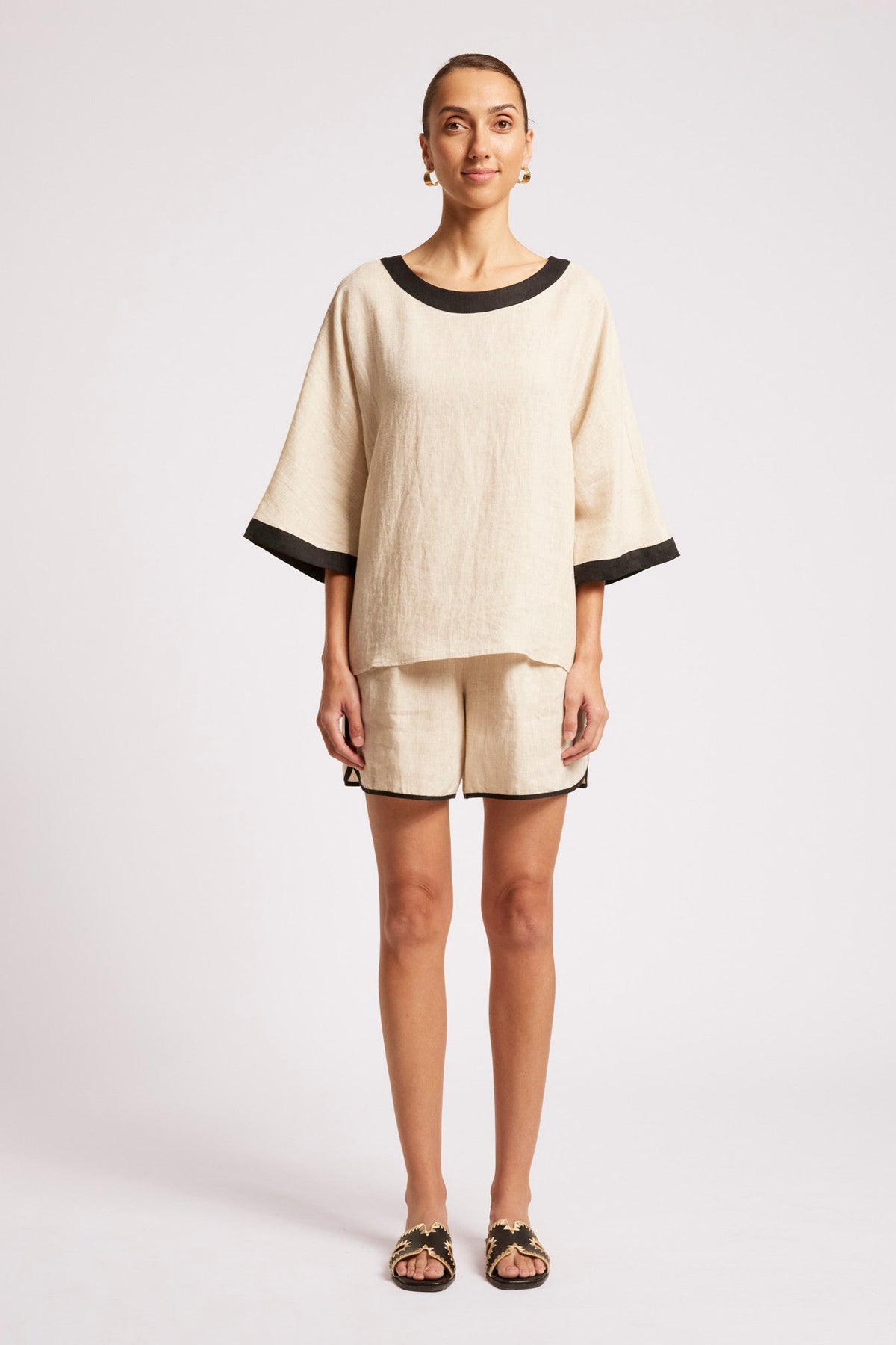 A woman modeling a matching two-piece linen set consisting of a short-sleeve top with black trim and matching shorts, photographed against a light gray studio background.