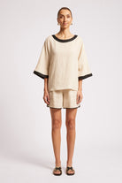A woman modeling a matching two-piece linen set consisting of a short-sleeve top with black trim and matching shorts, photographed against a light gray studio background.