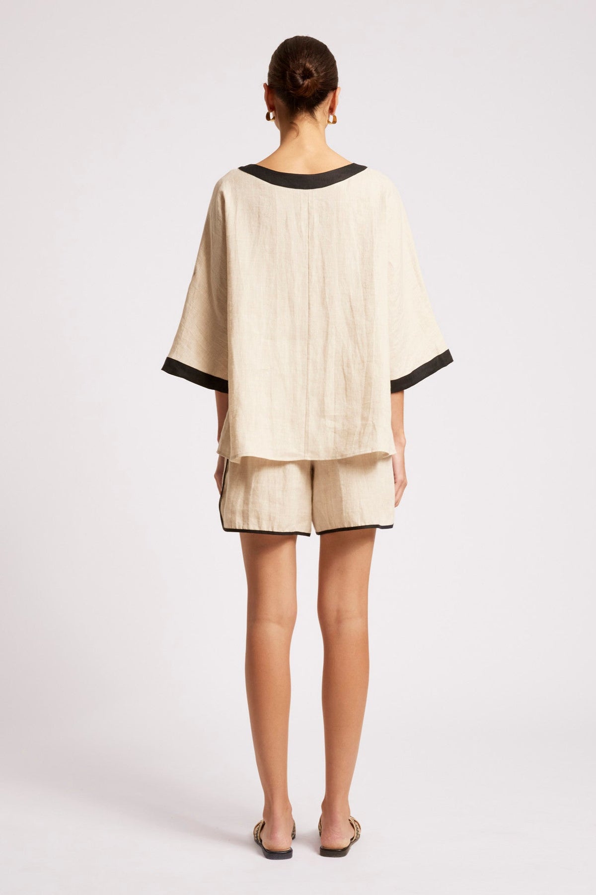 A woman viewed from the back wearing a light beige linen two-piece set consisting of a short-sleeve top with black trim and matching shorts, set against a plain light gray studio background.