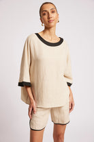 A woman modeling a matching beige linen lounge set with black trim, featuring a loose short-sleeve top and coordinating shorts, against a light gray studio background.