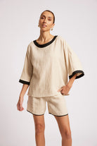 A woman modeling a light beige linen two-piece lounge set with black trim, featuring a short-sleeve top with elbow-length sleeves and matching shorts, shot against a plain light gray studio background.