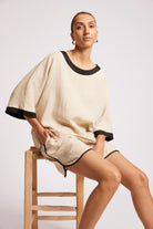 A woman seated on a wooden stool wearing a loose beige linen-like short dress or tunic with black trim at the neckline, sleeve cuffs, and hem, against a light gray studio background.