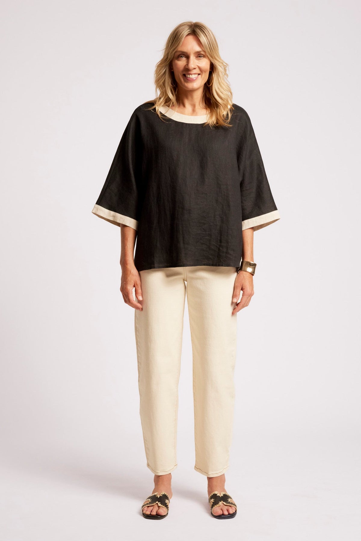 A woman modeling a black short-sleeve top with cream trim and cream pants against a light gray studio background.