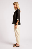A woman stands in profile wearing a loose black top with rolled three-quarter sleeves and cream cuffs, paired with cream pants and black slide sandals, against a plain light gray studio background.