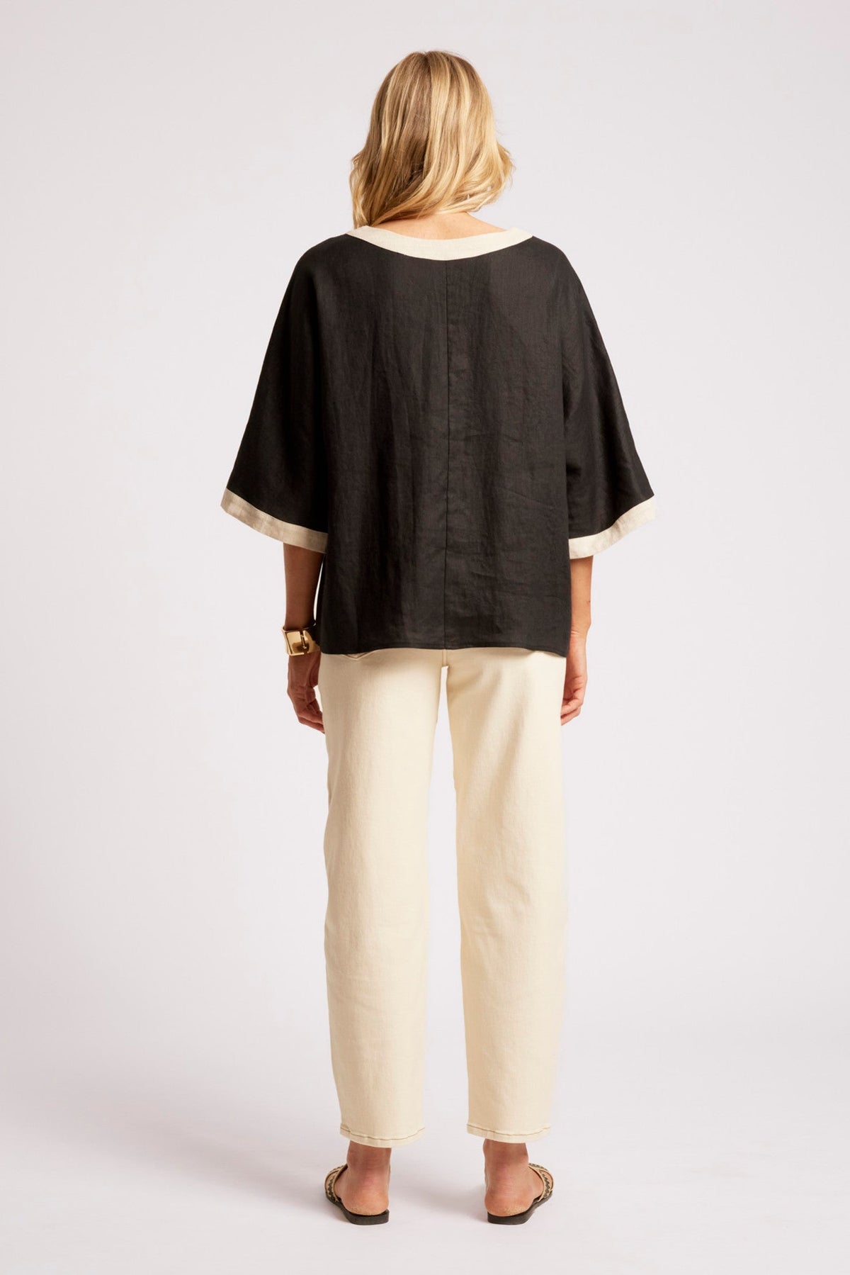 A woman shown from the back wearing a black short-sleeve top with cream trim and cream pants, standing against a light gray studio background.