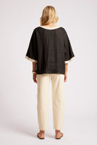 A woman shown from the back wearing a black short-sleeve top with cream trim and cream pants, standing against a light gray studio background.
