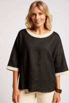 A woman wearing a black short-sleeve top with contrasting cream trim at the round neckline and sleeve cuffs, paired with cream pants, against a light gray studio background.