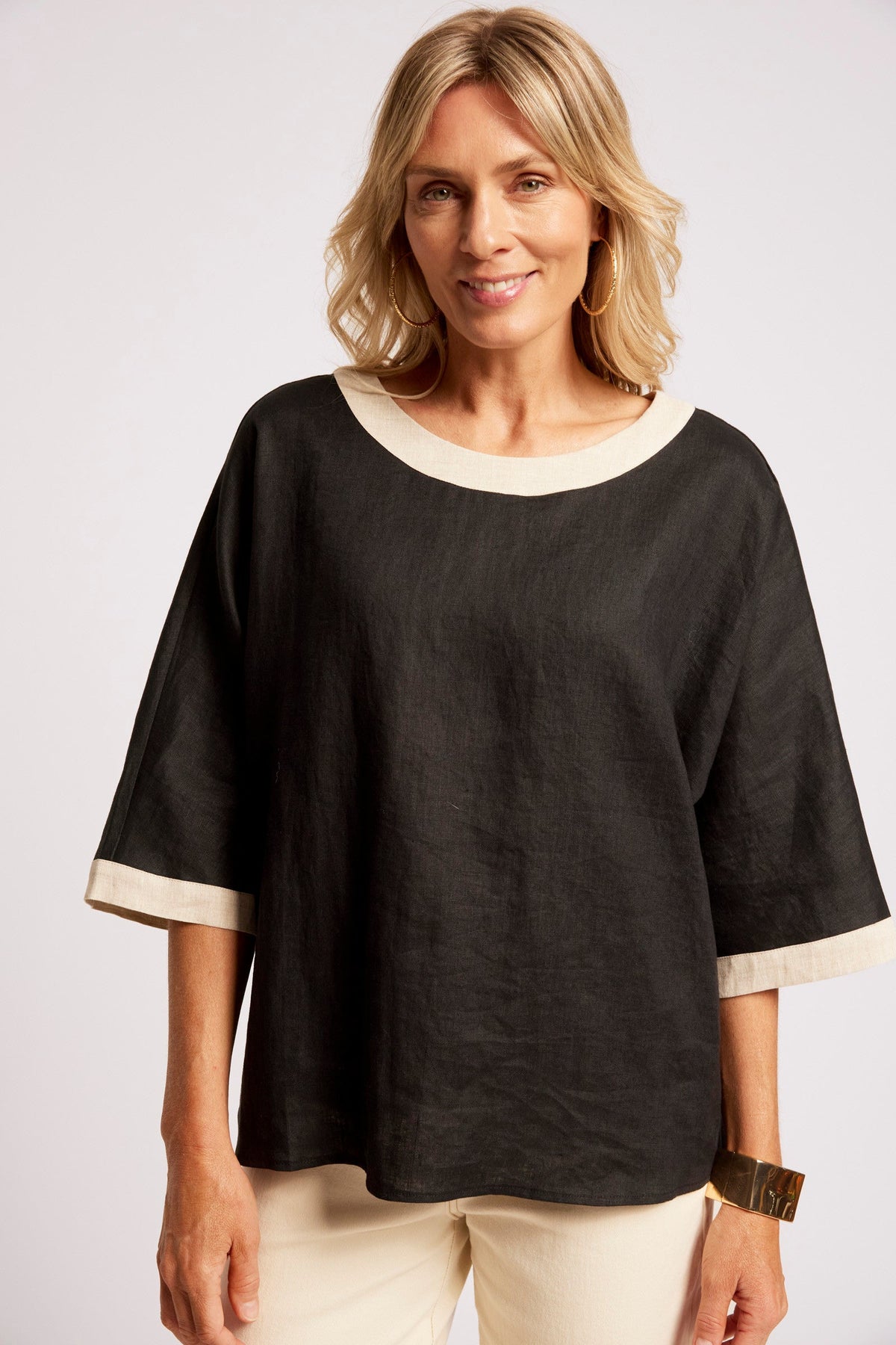 A woman wearing a black short-sleeve top with contrasting cream trim at the round neckline and sleeve cuffs, paired with cream pants, against a light gray studio background.