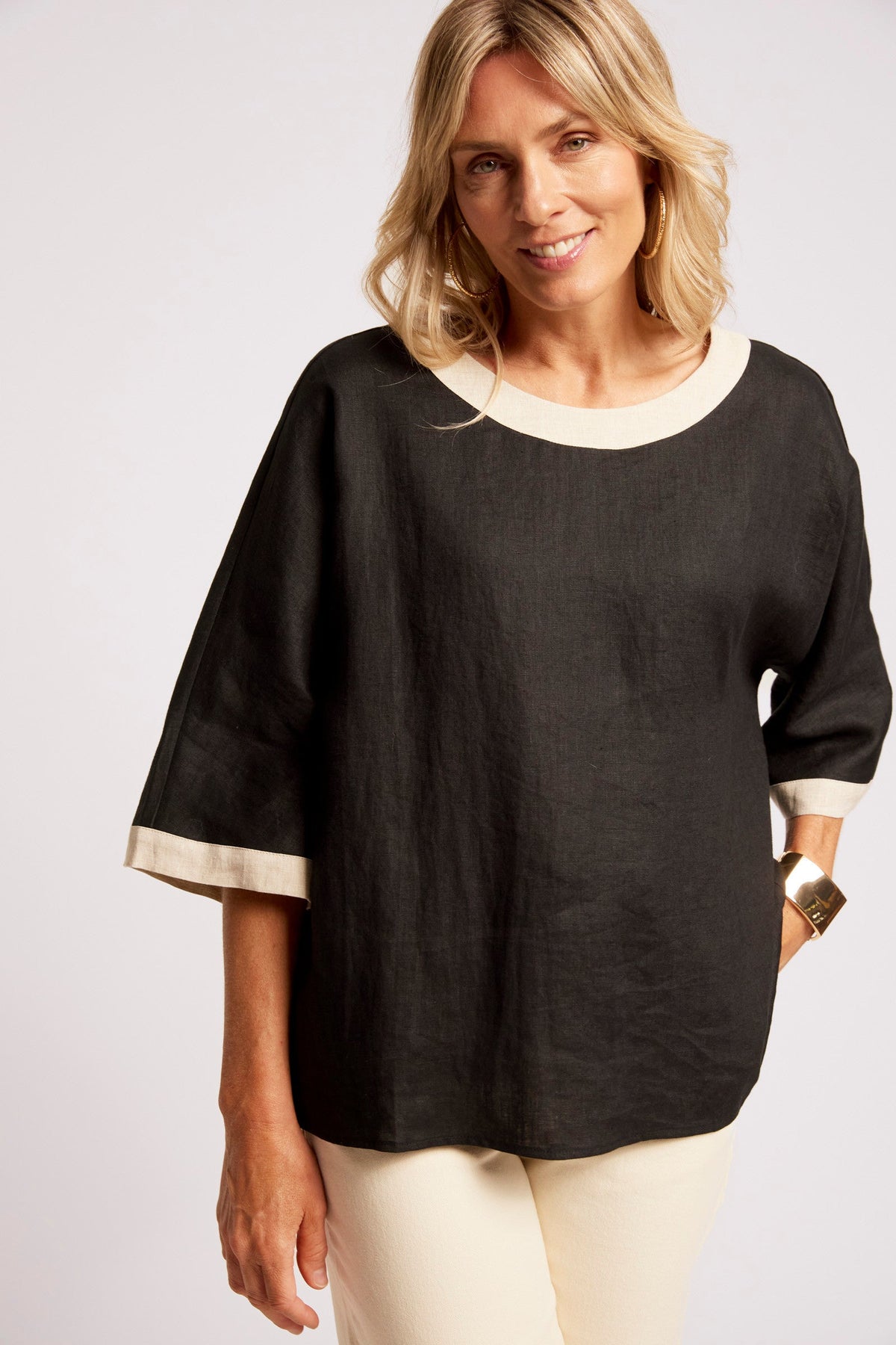A woman wearing a black linen-like top with cream trim at the round neckline and sleeve cuffs, paired with cream pants, photographed against a light gray studio background.