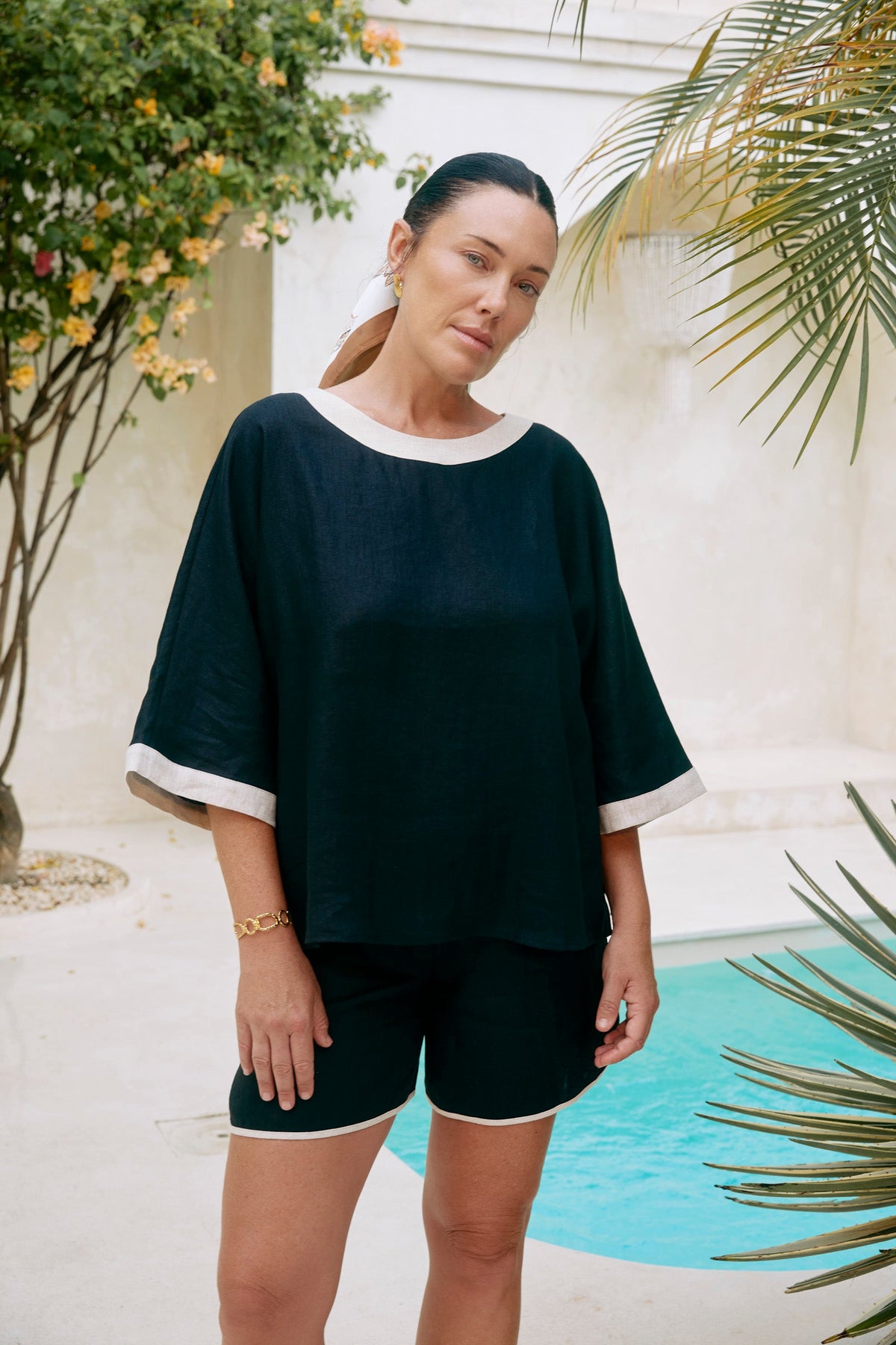 A woman wearing a black two-piece lounge set with white trim, standing outdoors next to a swimming pool with tropical plants.