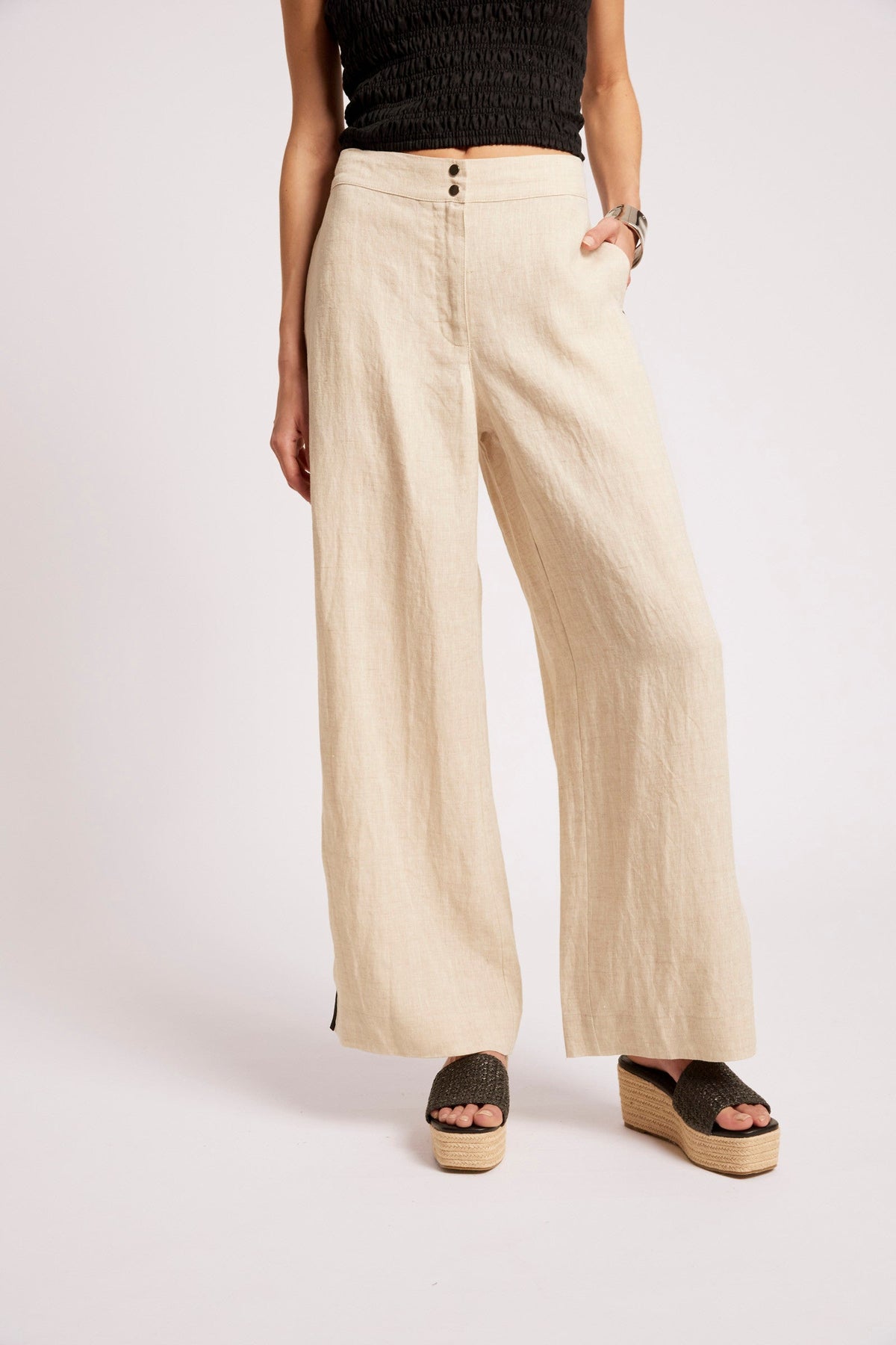 A model wearing high-waisted, wide-leg linen pants in a light beige color, paired with a black textured top and brown platform wedge sandals, against a plain light gray studio background.
