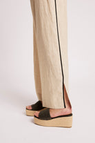 A close-up of a person's feet wearing beige wide-leg linen trousers with a black side stripe and black woven platform slide sandals with espadrille wedge soles, shown against a light gray studio background.