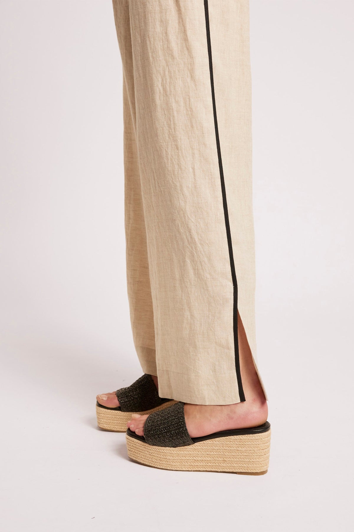 A close-up of a person's feet wearing beige wide-leg linen trousers with a black side stripe and black woven platform slide sandals with espadrille wedge soles, shown against a light gray studio background.
