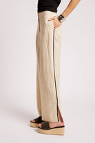 A person wearing wide-leg beige trousers with a thin black side stripe, paired with black platform slide sandals, shown from the waist down against a plain light background.