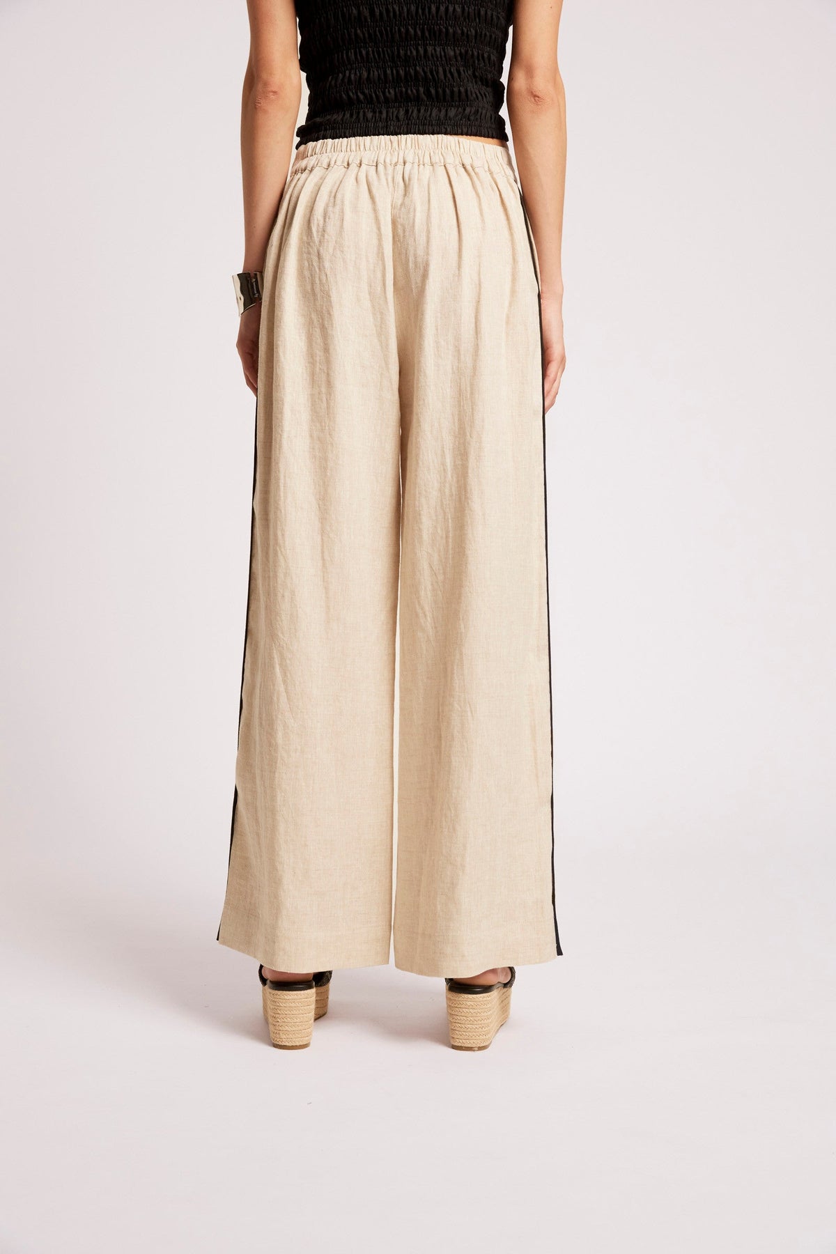 A model is shown from the back wearing wide-leg linen trousers in a light beige color with black side stripes, paired with a black top and beige platform sandals.